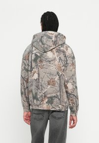 Kapuci sporta krekls ar kamuflāžas rakstu, kas ietver pelēkas, brūnas un zaļas toņus. Mīksts audums ar brīvu piegriezumu un ribotas piedurknes.