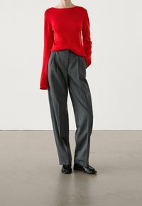 Un pull en tricot rouge à manches longues, associé à un pantalon gris à taille haute et plissé. Des chaussures noires complètent l'ensemble.