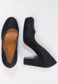 Zapatos de tacón alto negros con parte superior de tela texturizada y tacón cuadrado. El interior es marrón, con una suela estampada.