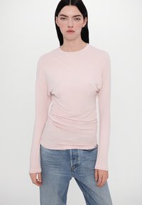 Light pink long-sleeve top with a round neckline, featuring a ruched texture on the front and straight sleeves, paired with blue jeans.