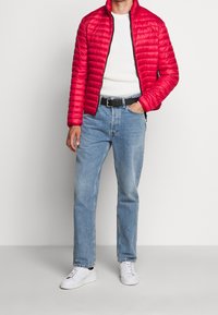 Červená пуховка s předním zipem a ribovanou strukturou, kombinovaná s bílým pleteným svetrem, světlými modrými džínami a bílými teniskami.