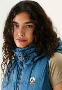 Chaqueta acolchada azul con collar alto, que presenta detalles de doble hebilla y una patch de logo, llevada sobre una blusa de color beige claro. Tela texturizada.