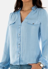 Blusa azzurro chiaro a maniche lunghe realizzata in tessuto morbido, caratterizzata da due tasche sul petto, bottoni argentati e polsini arricciati. Design aderente.