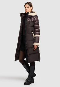 Cappotto imbottito lungo di colore marrone scuro con cappuccio foderato in pile crema, chiusura frontale con zip, tasche laterali e polsini in pile a contrasto. Indossato con stivali alti neri.