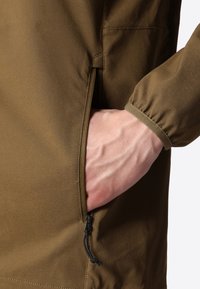 Brun jakke med sidelomme, der har en blød tekstur og elastiske manchetter. Snøre detaljer ved lommen forbedrer funktionaliteten.