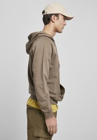 Felpa marrone con tasca a marsupio, cappuccio con lacci, indossata sopra una maglietta gialla. Un cappello beige e pantaloni cargo verde oliva completano l'outfit.