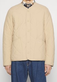 Beige Steppjacke mit rundem Ausschnitt, Druckknopfverschluss und Vordertaschen. Leichter Stoff mit subtiler Textur.