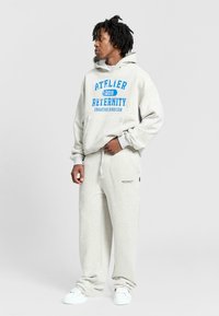 Jasnoszary zestaw bluzy z kapturem i spodni dresowych z niebieskim nadrukowanym napisem oraz elastycznym paskiem. Miękka, teksturowana tkanina i białe sneakersy dopełniają całość.