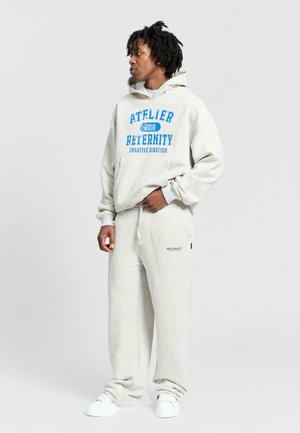 Completo in felpa grigio chiaro composto da hoodie e pantaloni della tuta, caratterizzato da testo stampato blu e una vita con cordino. Il look è completato da un tessuto morbido e strutturato e sneakers bianche.