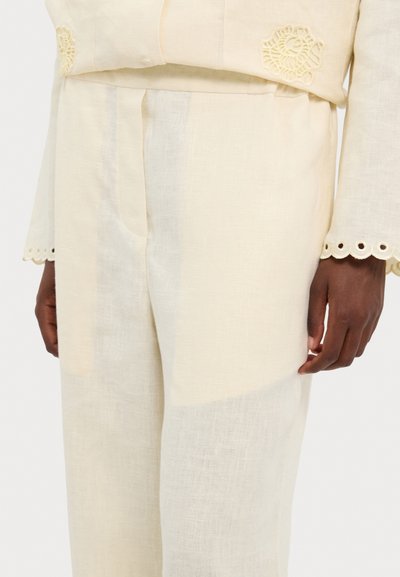 Tenue en lin couleur crème avec un tissu texturé. Présente une broderie complexe sur le haut et des bords festonnés décoratifs sur les manches.