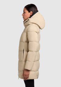 Woman in profile wearing a beige hooded puffer coat with black zipper details and black pants against a plain gray background.