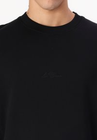 Zwarte sweatshirt met een geribbelde halslijn. Beschikt over een subtiel, reliëflogo in het midden. Gemaakt van een zachte, rekbare stof.