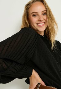 Blusa negra con mangas plisadas y texturizadas y un diseño de botones. Presenta pequeños puntos elevados y un intrincado detalle en el collar.