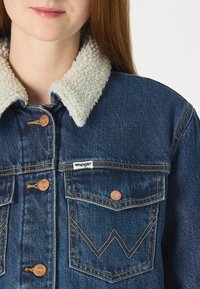 Τζάκετ από τζιν με ελαφρύ, αφράτο κολάρο σε κρέμα απόχρωση, χάλκινα κουμπιά και λογότυπο Wrangler. Διαθέτει δύο θηκάκια στο στήθος και ραφές σε αντίθεση.