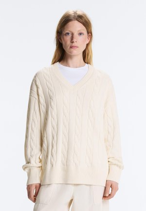 Kobieta z prostymi jasnobrązowymi włosami, ubrana w oversize kremowy sweter warkoczowy z dekoltem w serek oraz dopasowane jasne spodnie.