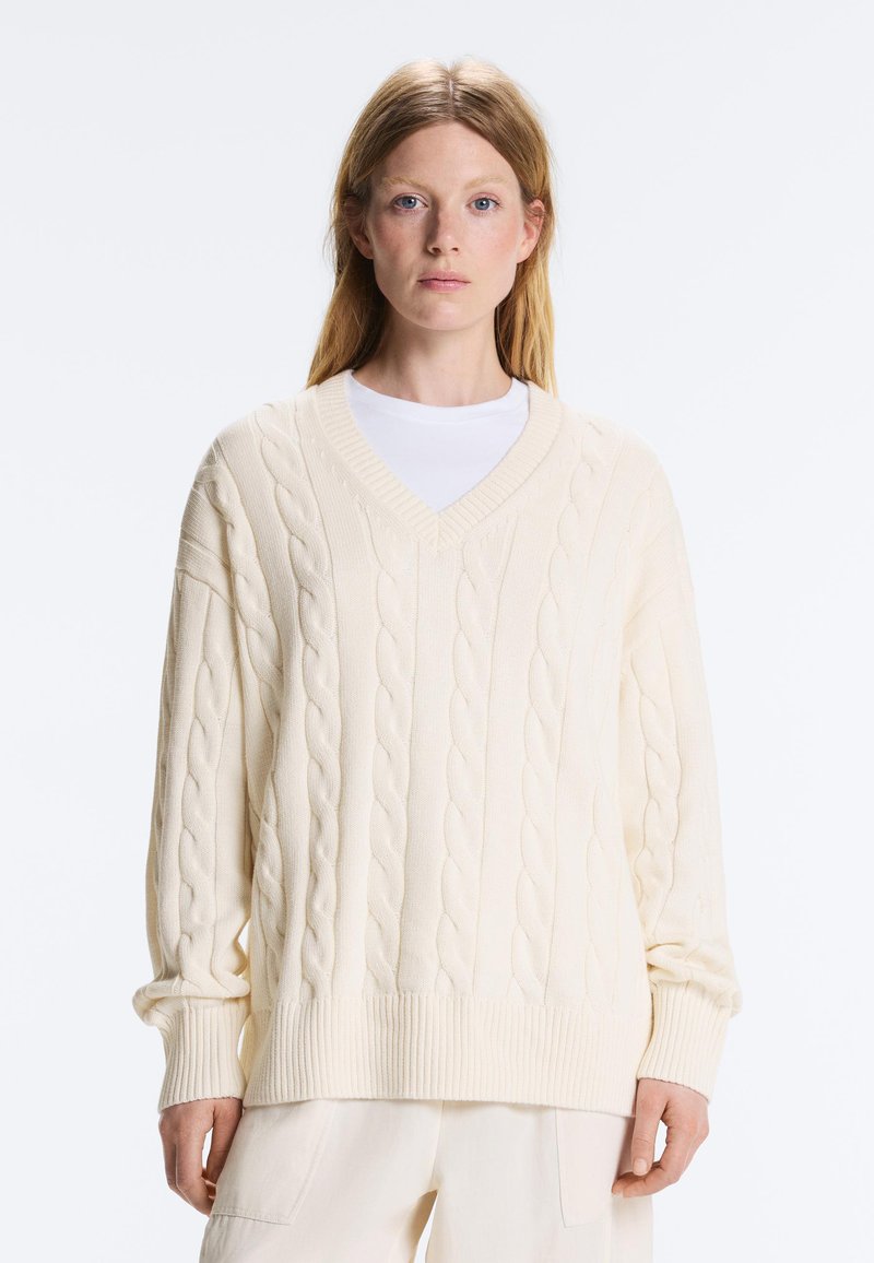 Femme aux cheveux lisses châtain clair portant un pull oversize crème en maille torsadée à col en V et un pantalon clair assorti.