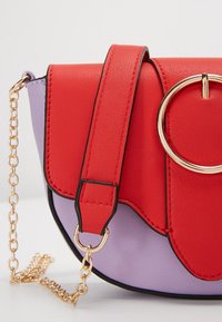 Červená a levandulová kožená crossbody taška se zlatým kulatým kovovým akcentem, vybavená texturovaným popruhem a vlnitým designem.