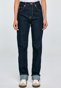 Jeans de mezclilla azul oscuro con una cintura ajustada, perneras rectas, puños enrollados y detalles de costura amarillos. Combinados con zapatillas de corte bajo.