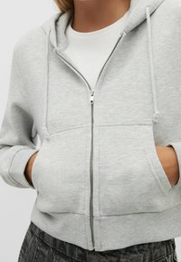 Sudadera con cremallera de color gris claro hecha de tela suave, con capucha ajustable, bolsillos frontales y puños acanalados. Diseño simple y casual.