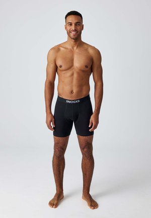 Uomo sorridente senza maglietta in piedi a piedi nudi che indossa boxer neri con fascia elastica "SNOCKS" su uno sfondo chiaro semplice.