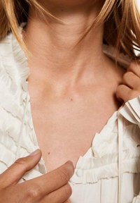 Witte blouse met rimpeldetail langs de halslijn, met een knoopsluiting. Gladde textuur en delicate stof met een subtiele glans.