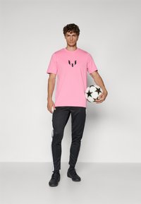 Pink short-sleeve t-shirt with black logo detail, paired with black tapered pants featuring white side stripes, worn with black athletic shoes.