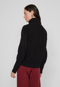 Melns adīts džemperis ar augstu apkakli, ribotu tekstūru un pieguļošu piedurkni. Kombinēts ar bordo biksēm, kas izceļ gaisīgas krāsas un brīvu piegriezumu.