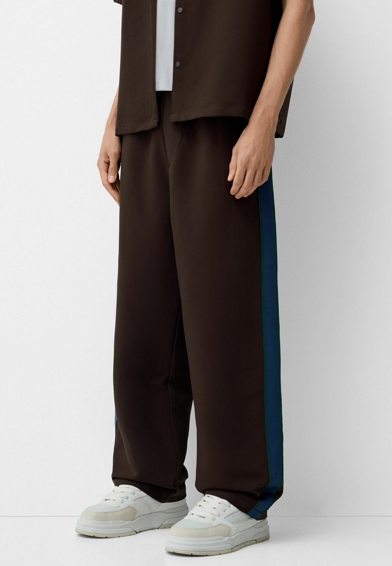 Pantalons larges marron avec des rayures latérales vertes et bleues, fabriqués à partir d'un matériau lisse. Associés à des baskets blanches pour le contraste.