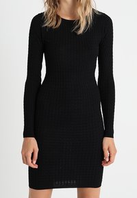 Robe noire en maille avec manches longues, silhouette ajustée et motif texturé. Col rond, longueur mi-cuisse, détails minimalistes.