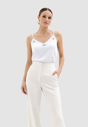 Donna che indossa una maglia bianca senza maniche con dettagli decorativi e pantaloni bianchi a vita alta, in piedi con una mano in tasca su uno sfondo grigio.