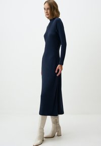 Marineblauw geribbeld langetermijn jurkje met een getailleerde bustier, een uitlopende midi-rok en een zijsplit, gecombineerd met witte kniehoge blokhaklaarzen.