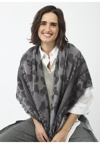 Écharpe grise avec motif léopard, bords à franges et texture tissée, drapée sur un pull léger et une chemise blanche, tenue par une personne.