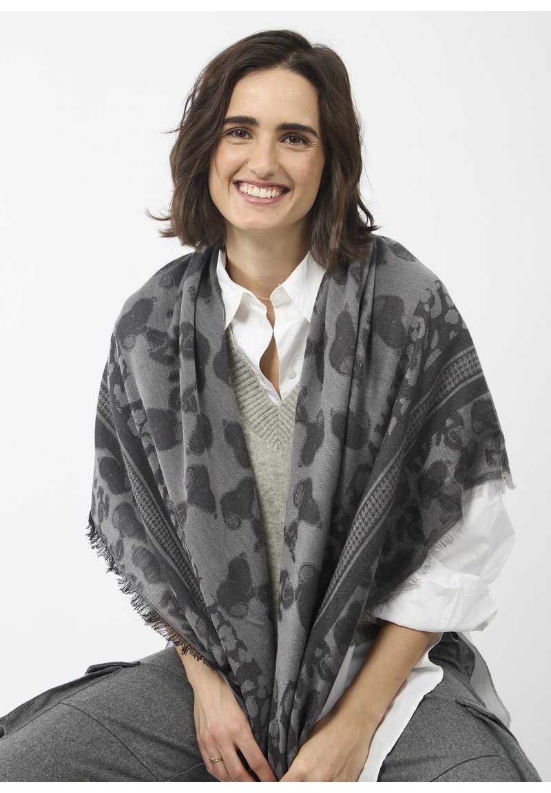 Écharpe grise avec motif léopard, bords à franges et texture tissée, drapée sur un pull léger et une chemise blanche, tenue par une personne.