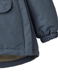Sininen eristetty takki, jossa on sileä ulkopinta, joustavat hihansuut ja pehmeä fleecvuoraus helmassa. Vinoraitaisuus on näkyvissä.