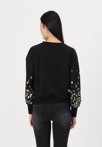 Svart hettegenser med lange ermer som har gull- og hvite blomsterdesign på ermene. Ribbestrikket nederkant og mansjetter, ledig passform.