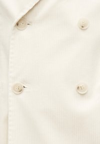 Κοντινό πλάνο λευκού σακακιού σεφ με διπλό πέτο, τέσσερα εμφανή κουμπιά και διακριτικές κάθετες ρίγες.