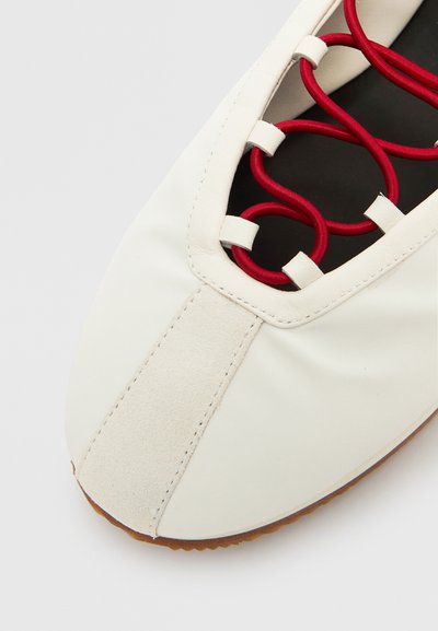Hvit skinnsko med rødt snøre som går gjennom utskjæringer, en semsket detalj ved tåen og rund form. Gummisåle.