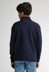 Maglione blu navy con un alto colletto, tessuto testurizzato e spacchi laterali. Abbinato a jeans azzurri chiari con dettagli di cucitura sulle tasche posteriori.