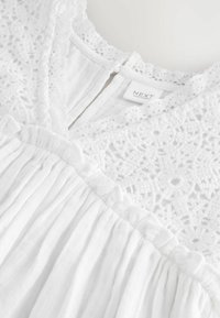 White dress featuring a textured lace bodice and a pleated fabric skirt with a ruffled accent along the seam. Tag visible near the neckline.
