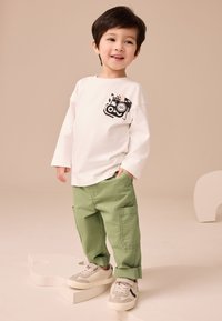 Chemise blanche à manches longues avec un motif noir sur la poche, associée à un pantalon cargo vert et des baskets aux tons neutres.