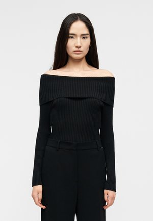 Kvinde med langt mørkt hår iført en tætsiddende sort ribstrikket sweater med bådudskæring og højtaljede sorte bukser, set forfra.