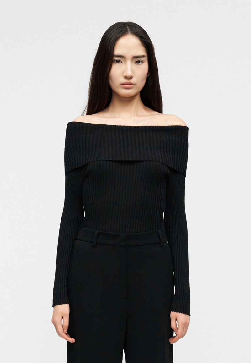 Γυναίκα με μακριά σκούρα μαλλιά, φορώντας στενό μαύρο ριμπ πλεκτό πουλόβερ χωρίς ώμους και ψηλόμεσο μαύρο παντελόνι, κοιτάζοντας μπροστά.