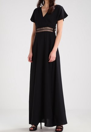Žena oblečená v černých maxi šatech s krátkými rukávy, výstřihem do V a háčkovaným detailem v pase, doplněná černými sandály na vysokém podpatku.