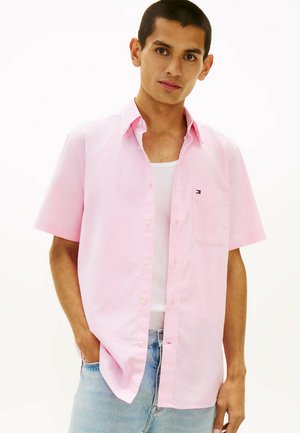 Junger Mann trägt ein hellrosa kurzärmeliges Hemd über einem weißen Tanktop und hellblauen Jeans, steht mit einer Hand in der Tasche.