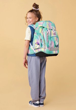 Lächendes Kind mit lockigem Haar zu einem Dutt gebunden, trägt einen großen pastellfarbenen Regenbogen-Rucksack mit Einhorn-Abzeichen, beigen Hemd, blauer Weste und grauer Hose.