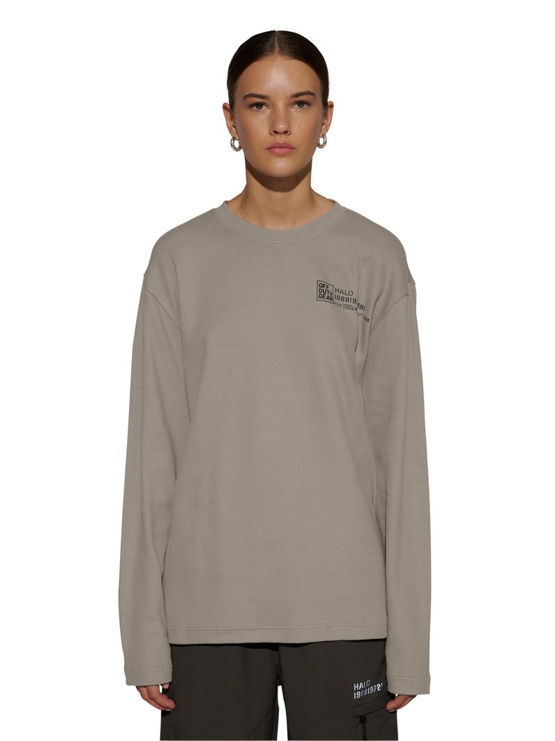 Frau trägt beige langärmliges Shirt mit kleinem schwarzem Text auf der Brust und dunkle Hose mit weißem Text am linken Oberschenkel, steht vor weißem Hintergrund.