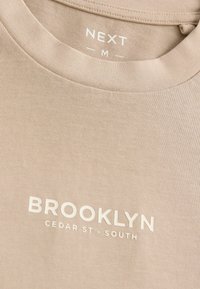 Beige bomulds T-shirt med rund halsudskæring, der har hvid tekst: "BROOKLYN CEDAR ST. - SOUTH" og mærket "NEXT M". Glat tekstur.