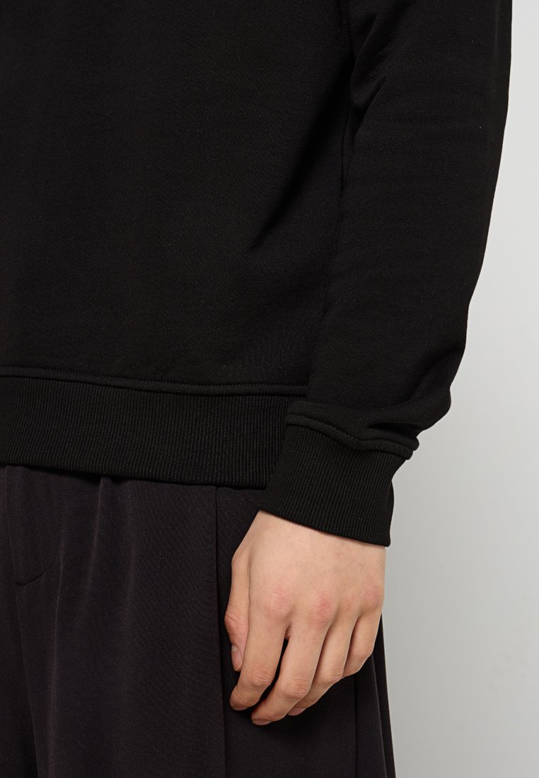 Sudadera negra con dobladillo y puños acanalados, con un corte relajado y textura suave, mostrando parcialmente una mano contra pantalones oscuros.