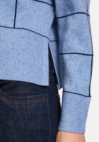 Hellblauer Pullover mit schwarzem Karomuster, geripptem Saum und Bündchen, seitlichen Schlitzen, kombiniert mit dunkelblauen Jeans.