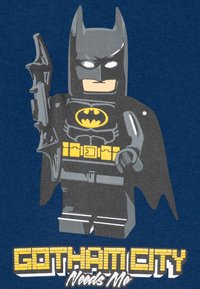 LEGO Batman-figur i svart kostym med cape, gul logotyp och bälte, som håller en grå gadget, på en marinblå bakgrund med texten "Gotham City Needs Me".